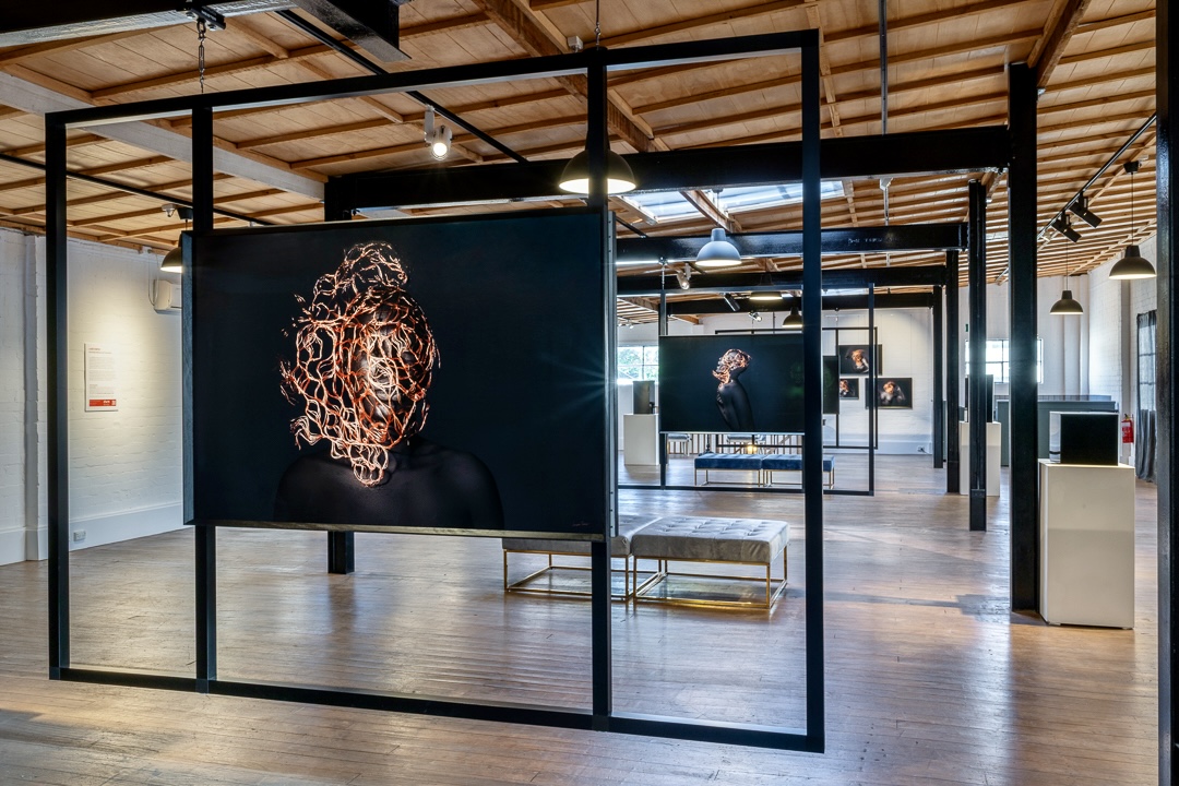 Free-standing black frame structure suspending a large portrait print in a gallery, part of the UNBECOMING exhibition